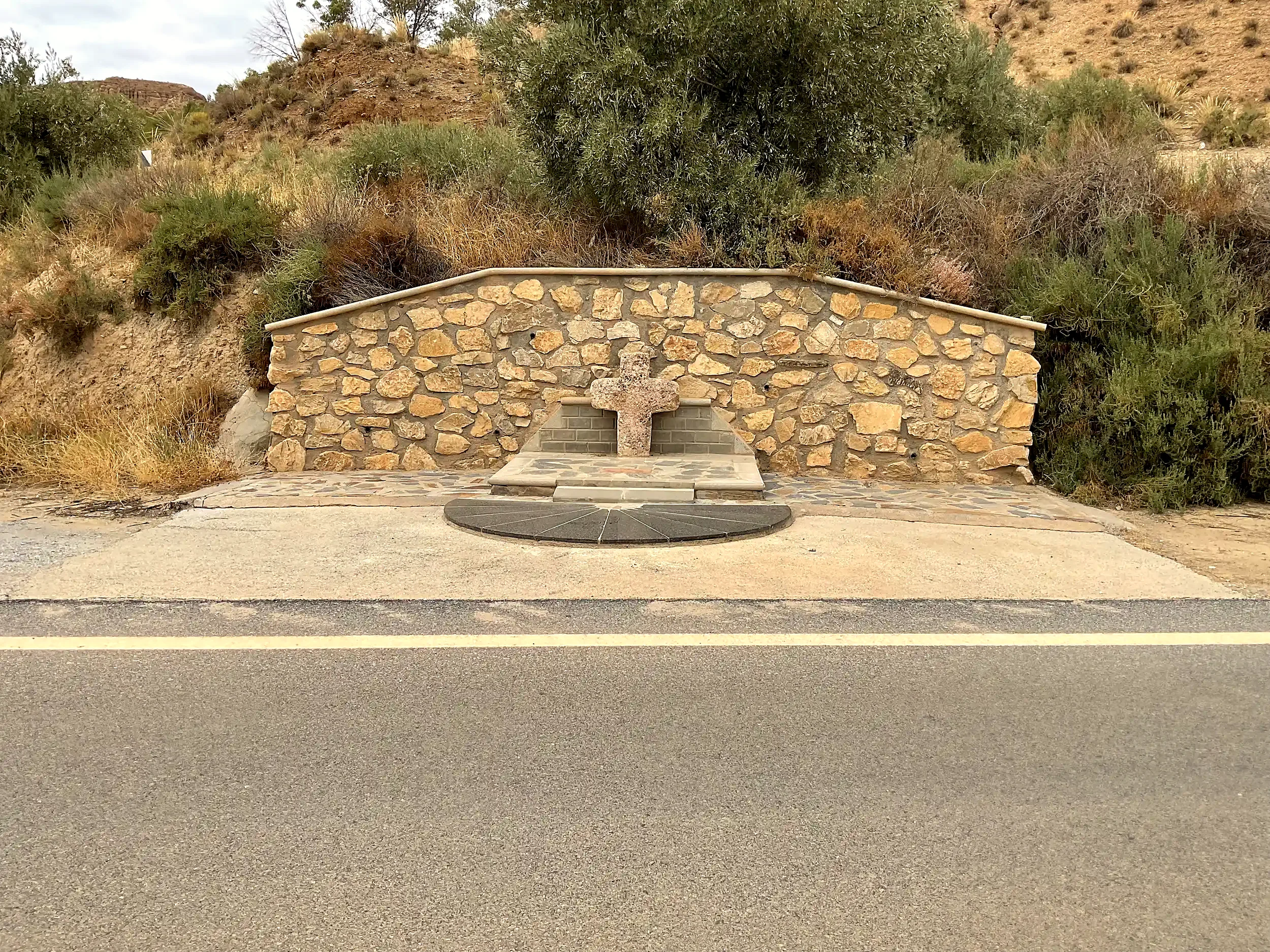 Vista frontal de la Cruz de San Marcos con muro de piedra, olivos y carretera